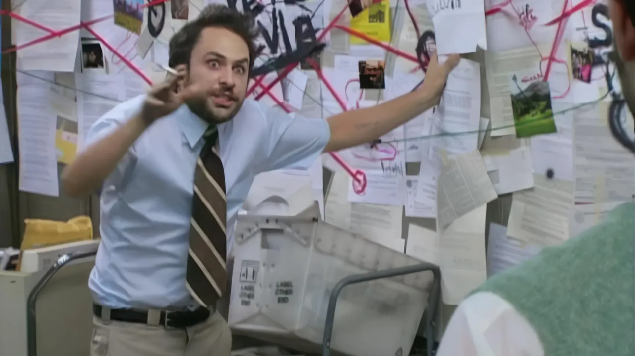 Man in office with disorganized bulletin board covered in papers and photos, appearing angry or confrontational, pointing and speaking to another person.