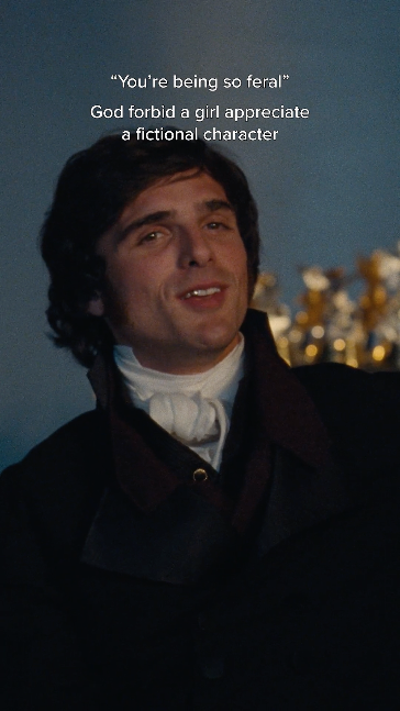 A man with dark hair and a slight smile, wearing a black coat and white shirt with a cravat, standing outdoors at night with blurred lights in the background. Text on the image reads: "You're being so feral" and "God forbid a girl appreciate a fictional character."