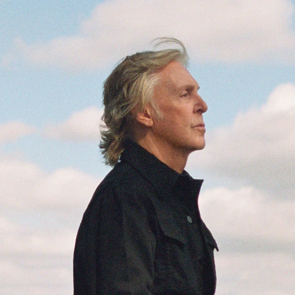 Profile view of a man with long gray hair, wearing a black jacket, against a cloudy sky.