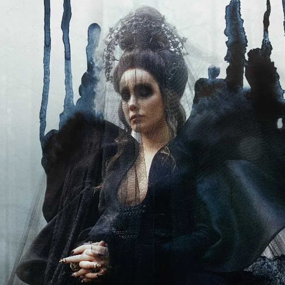 A woman with dark eye makeup and big hair in a high bun, wearing dark clothing, standing behind a glass surface with a distorted, abstract reflection.