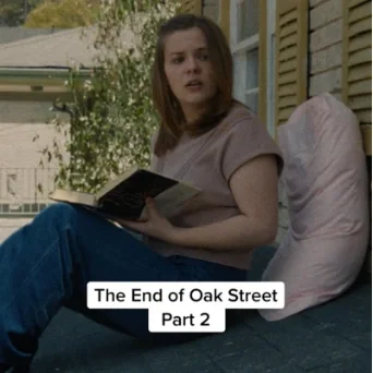 A young woman sitting outside on a porch, reading a book, with a pillow beside her. She is wearing a purple shirt and blue jeans.