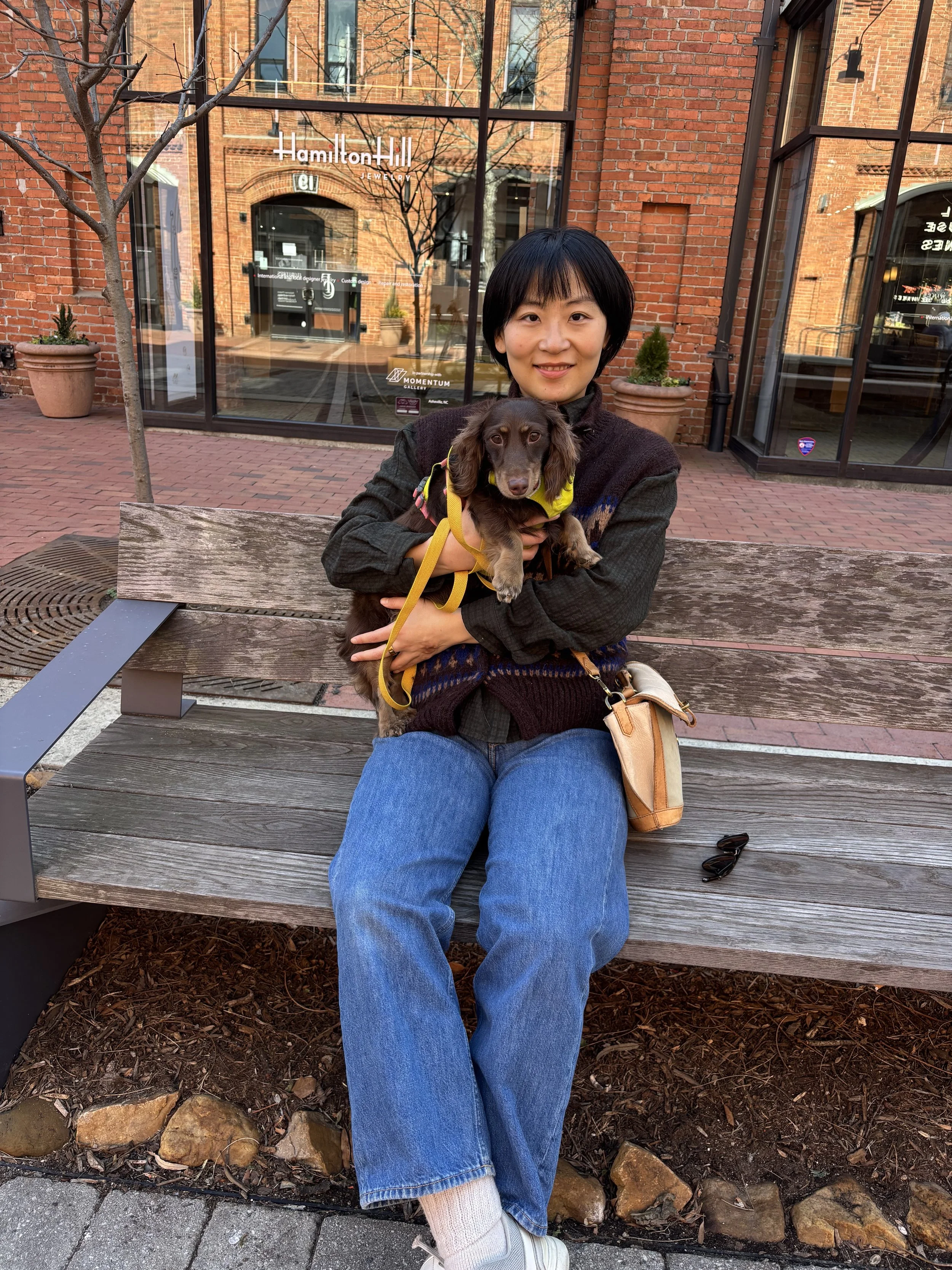 A woman sitting on a wooden bench outdoors, holding a brown dachshund dog in her arms. She is smiling and has short black hair, wearing a dark sweater and blue jeans. The background features a brick building with large glass windows and some potted plants.