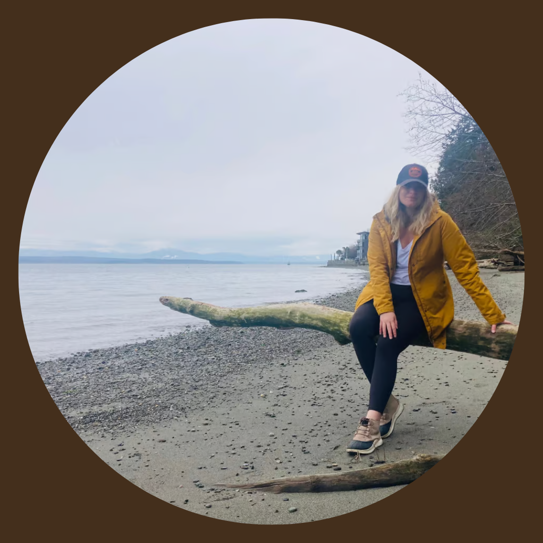 Elyssa wearing a yellow jacket, black leggings, and hiking boots sitting on a log at the beach with overcast skies, a calm lake, and trees in the background.