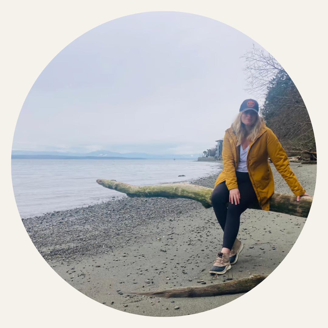 Elyssa sitting on a large piece of driftwood on a sandy beach, wearing a yellow jacket, black leggings, beige hiking boots, and a black cap, with trees and water in the background.