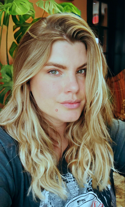 Elyssa with long wavy blonde hair indoors, with green plants and dark furniture in the background.