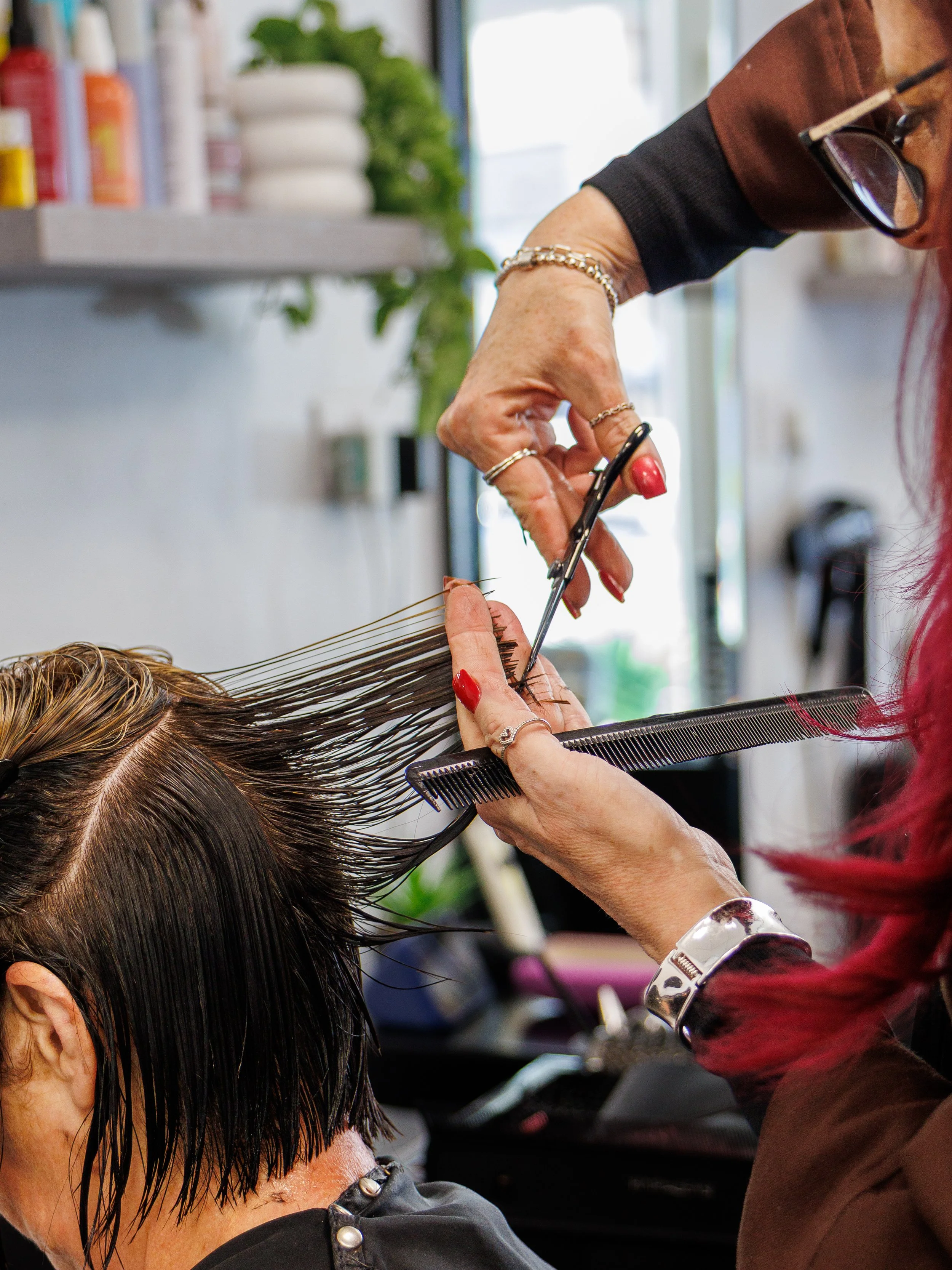 Hairdresser trims a client's wet, dark brown hair with scissors while holding a black comb at a salon.