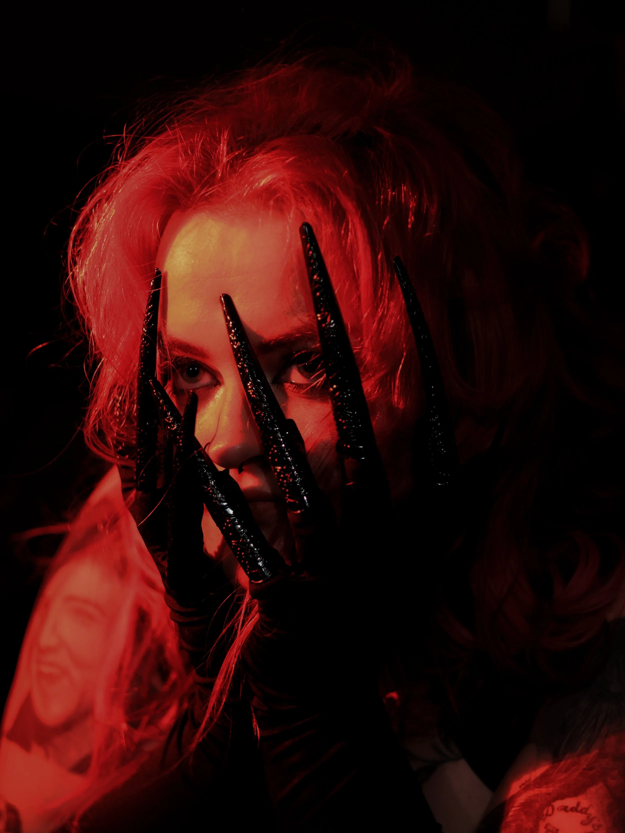 A woman with red hair holds her hand in front of her face with black, shiny, claw-like nails. The lighting is dark with a red glow.
