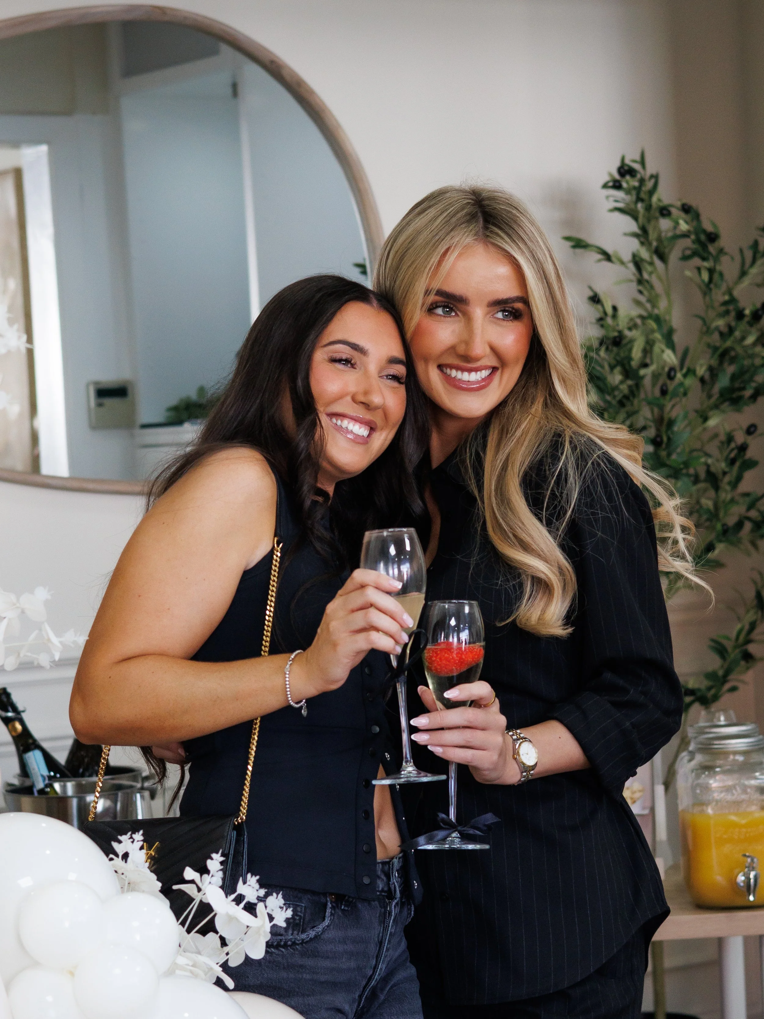 Two women smiling and posing together at a celebration, holding champagne glasses. One woman has dark hair and the other has long blonde hair. There are white balloons and flowers in the foreground.