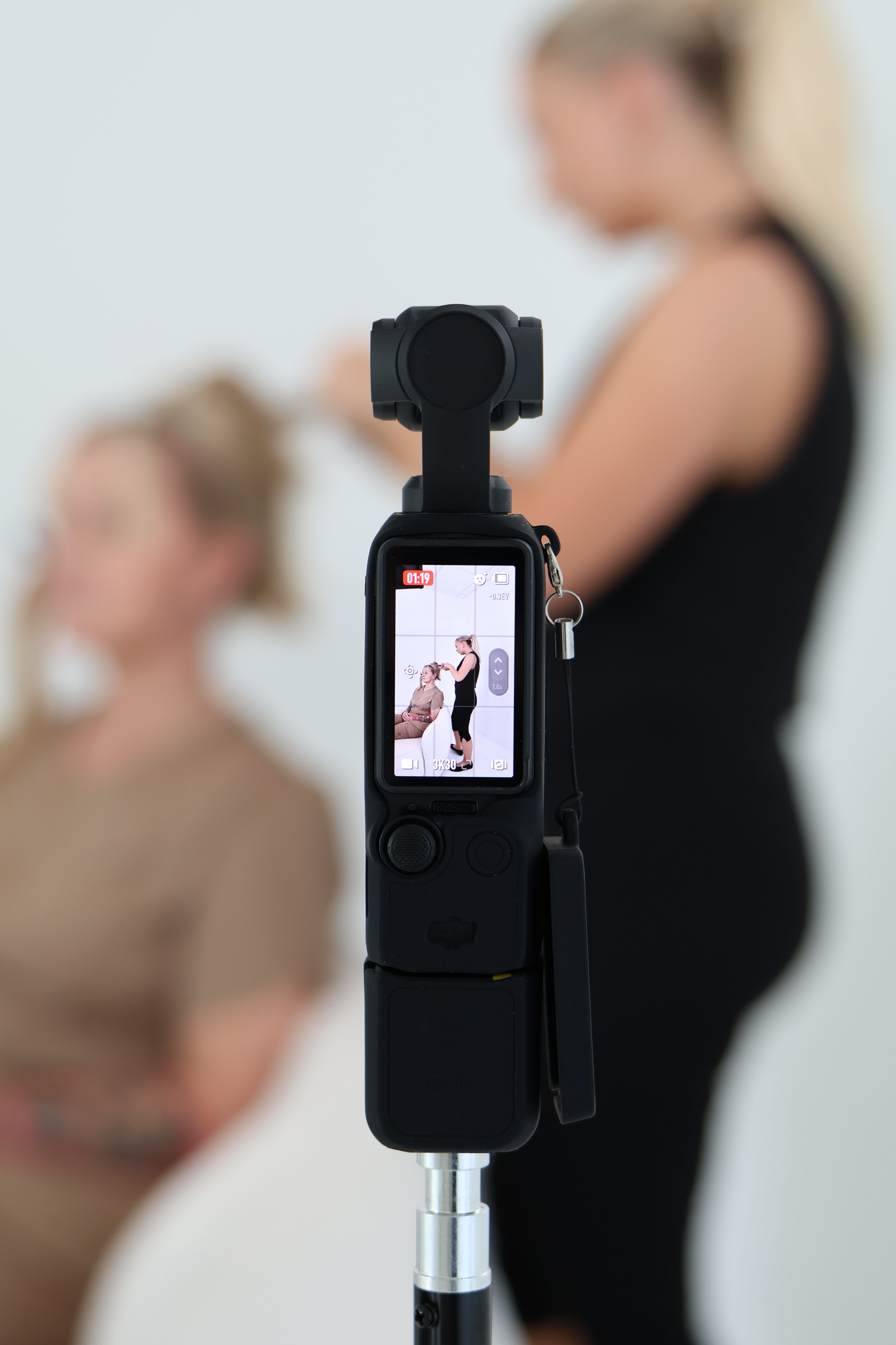 A camera on a tripod is recording two women, one sitting and the other standing and styling her hair, in a studio setting with a white background.