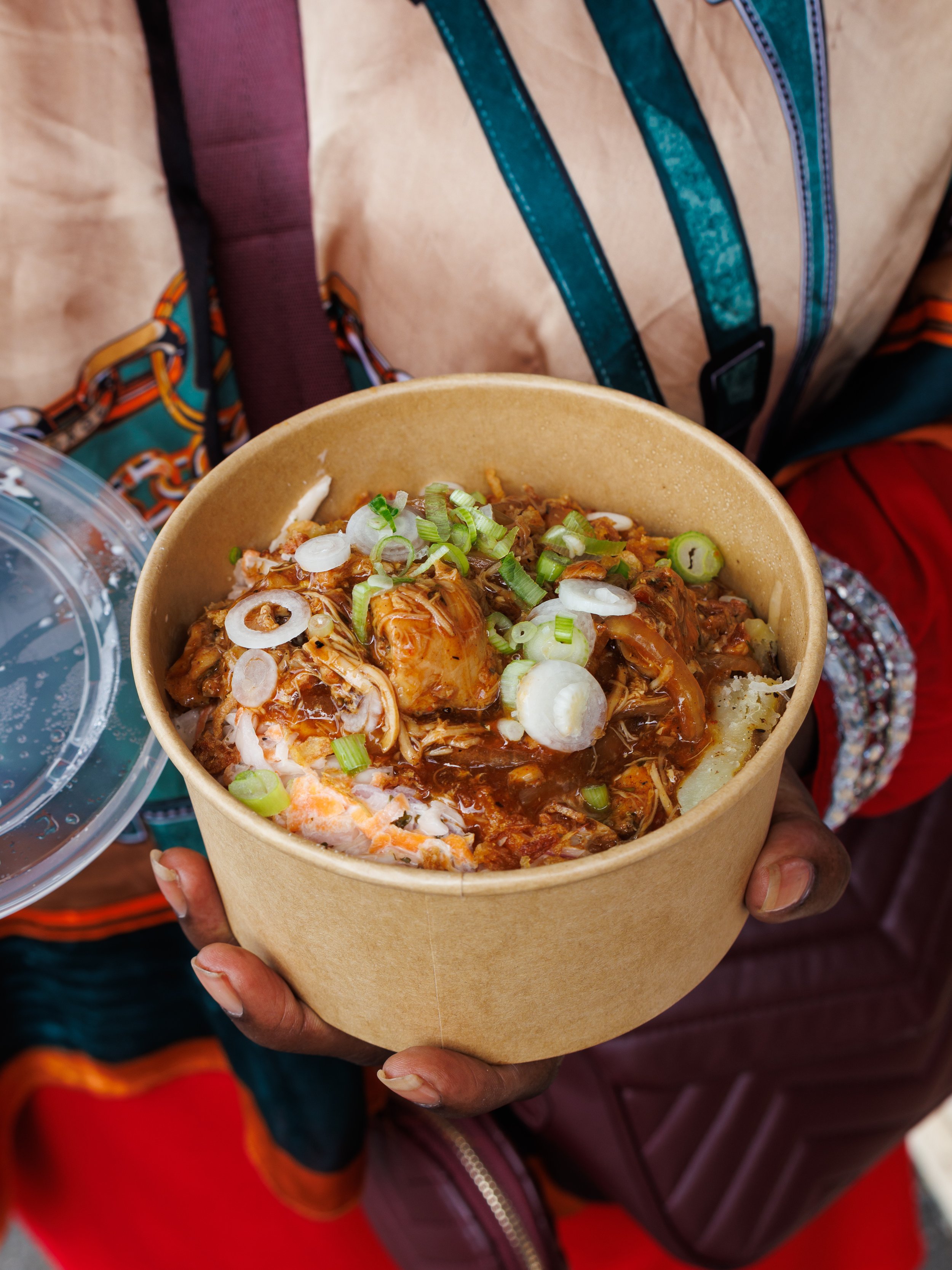 A person holding a brown paper bowl with a jacket potato topped with shredded meat and chopped spring onions.