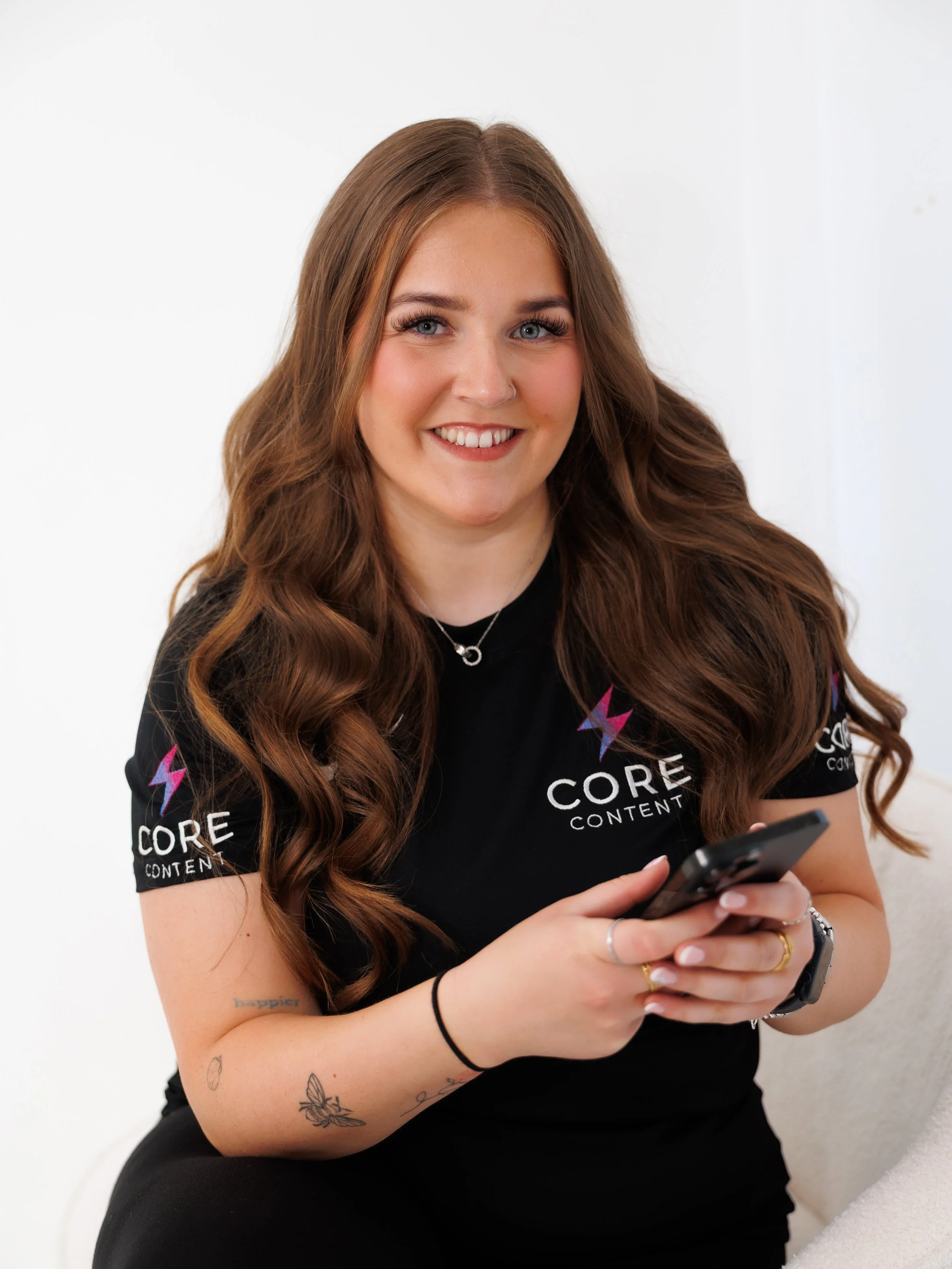 A young woman with long, wavy brown hair and blue eyes, smiling while looking at her phone. She has tattoos on her left arm and is wearing a black shirt with colorful logos that say 'CORE CONTENT' and features lightning bolts. She is sitting against a white background.