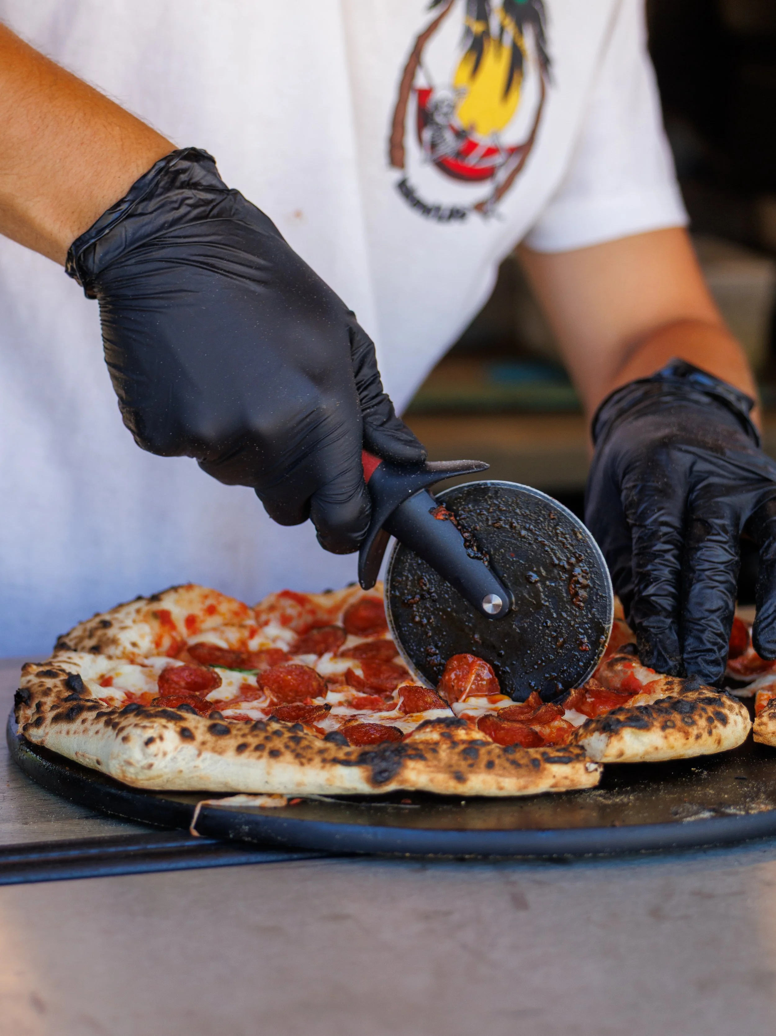 Person wearing black gloves slicing a cooked pepperoni pizza with a pizza cutter.