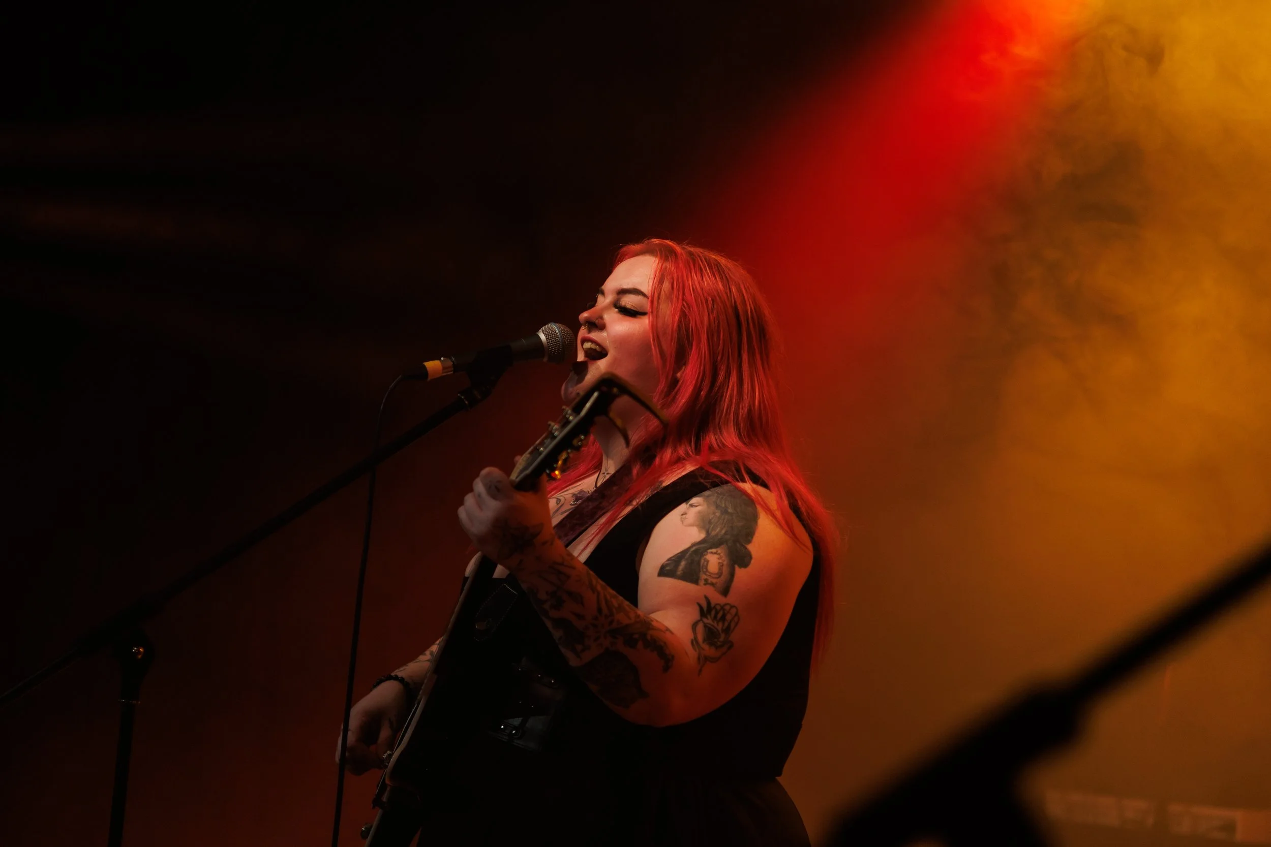 A woman with red hair singing into a microphone on stage, with tattoos on her arms and shoulders, illuminated by warm stage lights.