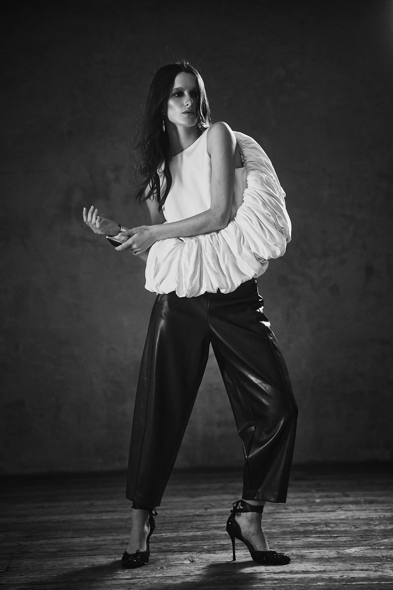A black and white photo of a woman standing on a wooden stage, wearing high-heeled shoes, a sleeveless top, and loose-fitting pants. Her dark hair is styled in loose waves, and she poses with one hand near her face and the other holding an object, against a plain dark background.