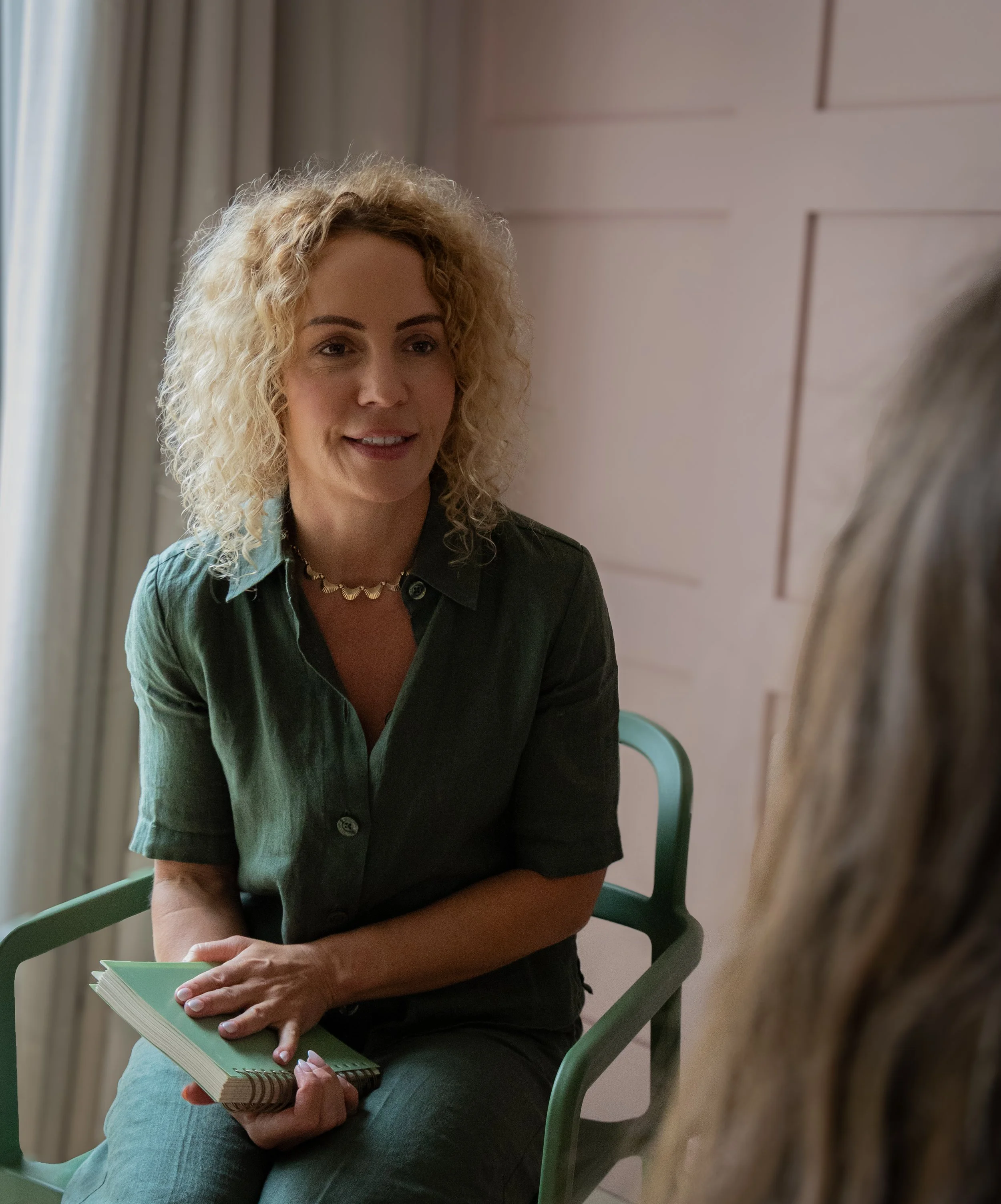 A woman with curly blonde hair sitting on a green chair, holding a notebook and a pen, in a room with beige walls and light-colored curtains.