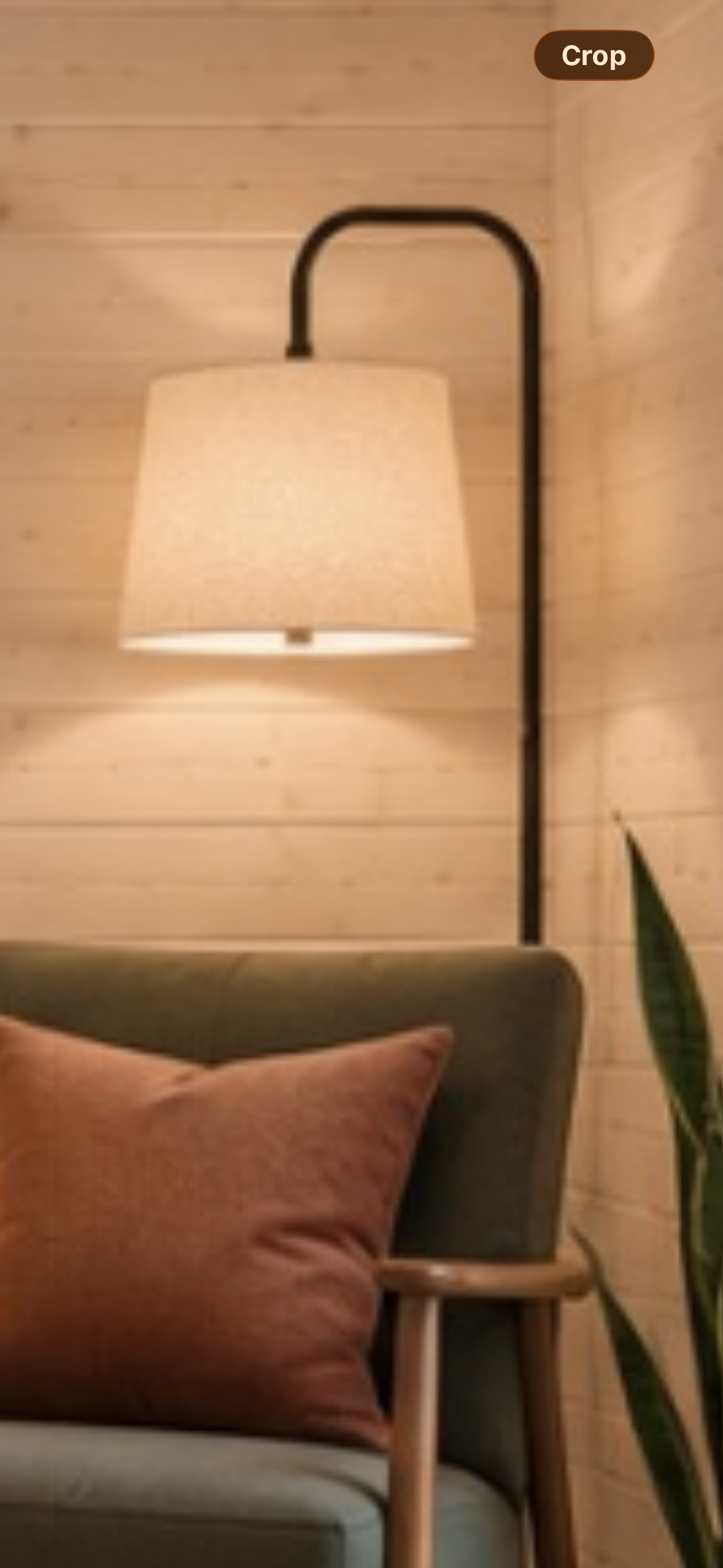 Wall-mounted lamp with a beige shade, a gray armchair with a brown pillow, wooden armrest, and a green plant in a cozy room with wooden walls.