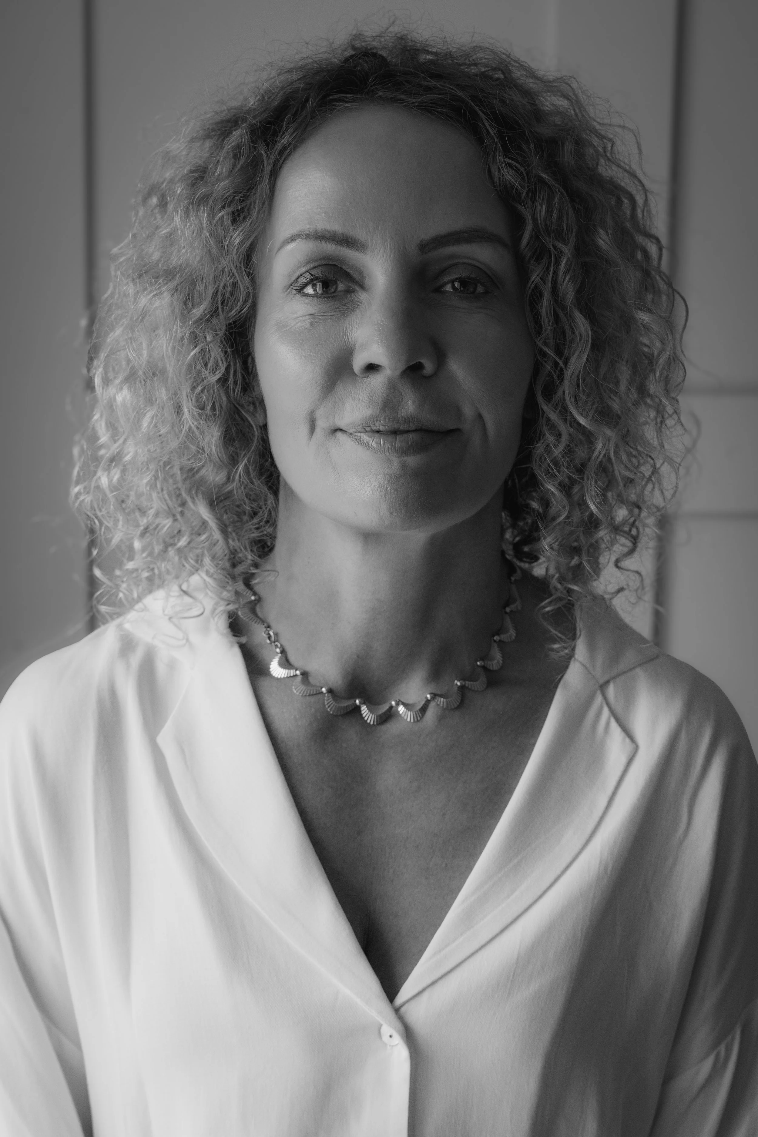 A woman with curly hair, wearing a necklace and a blouse, poses for a portrait in black and white.