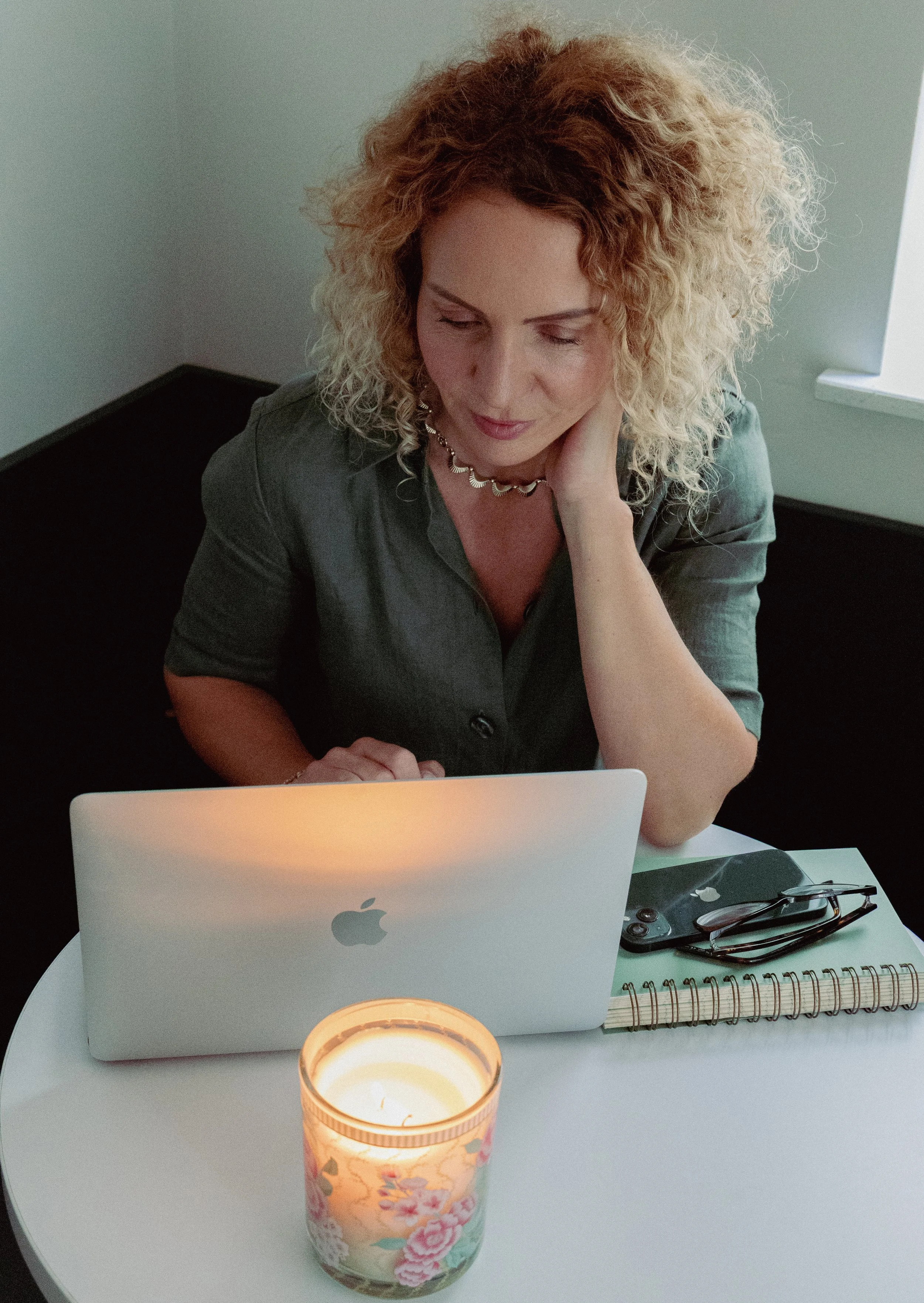 A woman with curly blonde hair sitting at a white table with a silver MacBook and glasses, with a closed notebook, a candle, and a smartphone nearby