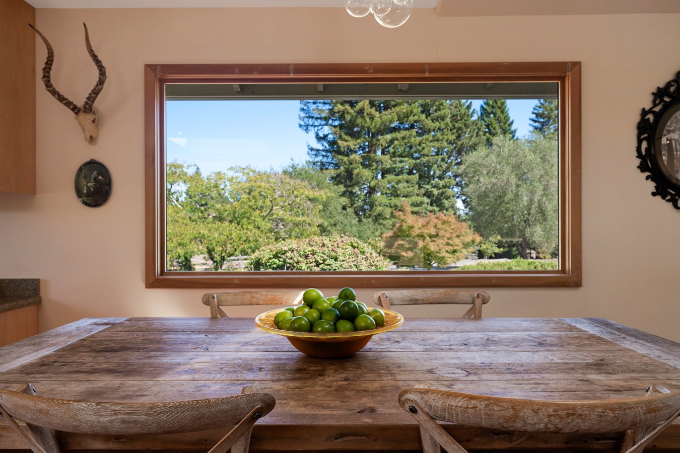 A wooden dining table with a bowl of green limes, located in a room with a large window showing a view of green trees outside.