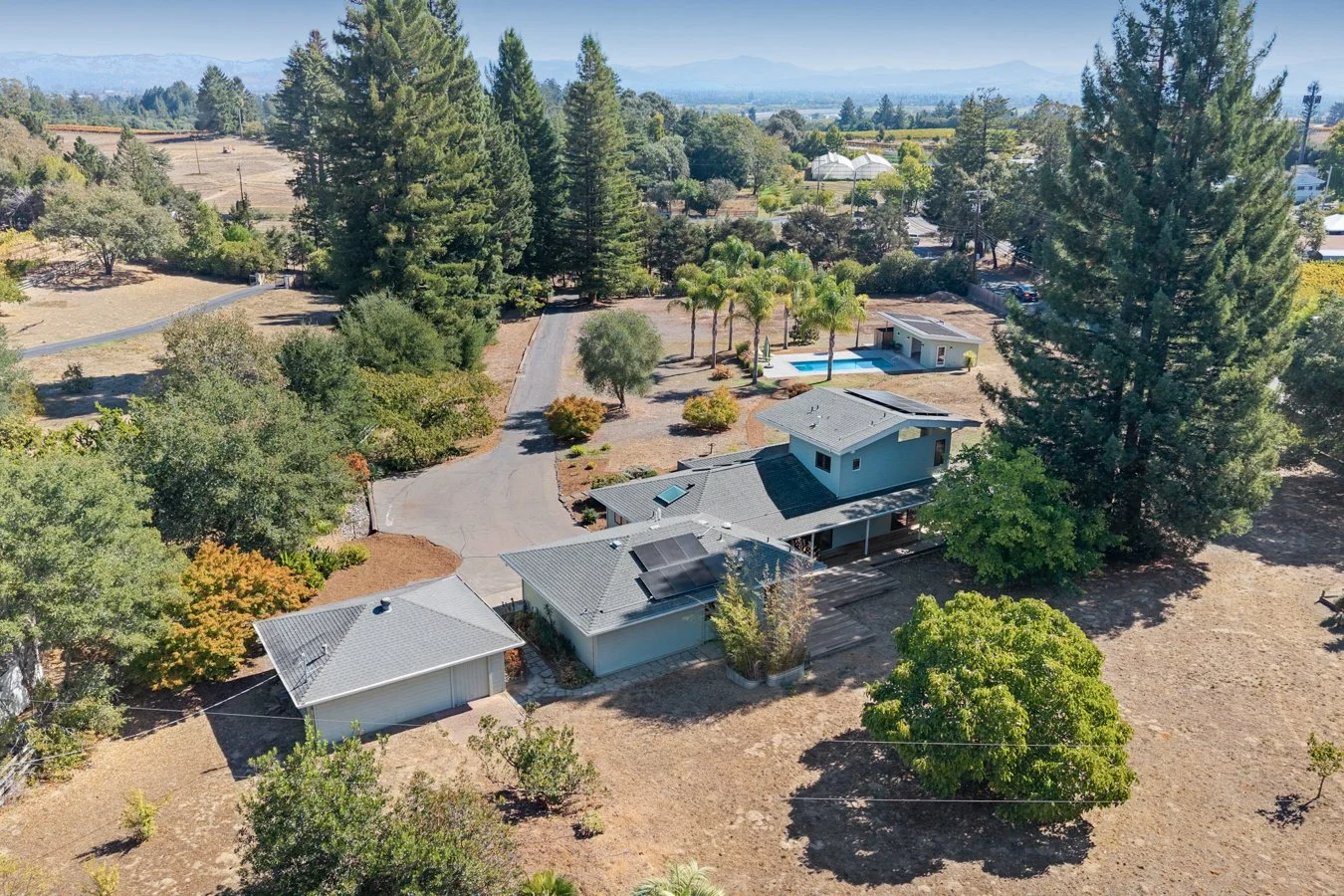 Aerial view of a residential property with a house, a small building with solar panels, a swimming pool, and surrounding trees in a rural area.