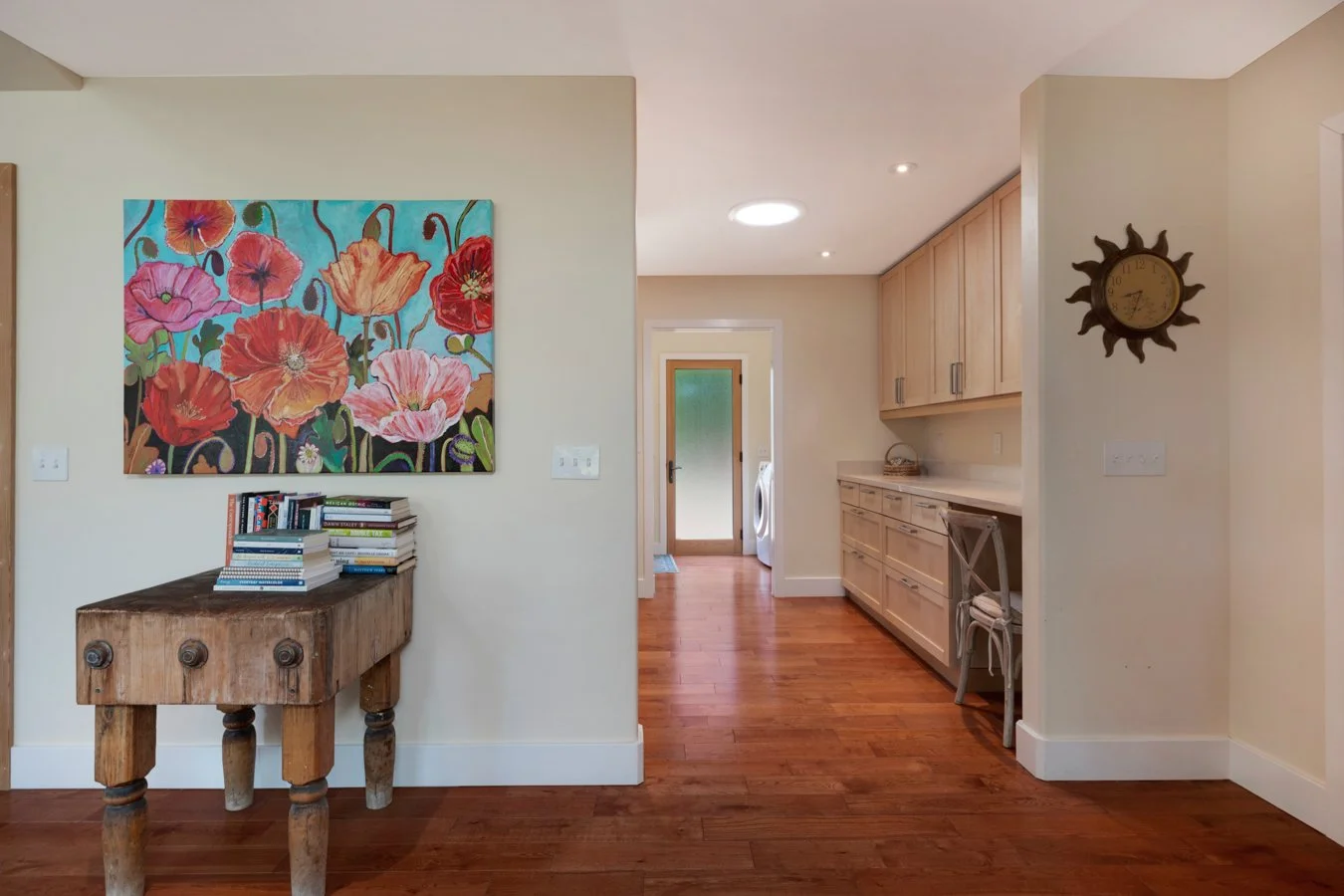 Interior of a home featuring a colorful floral painting on a cream wall, a rustic wooden table with books, and a kitchen area with light wood cabinets, a small chair, and a sun-shaped clock, with hardwood flooring throughout.
