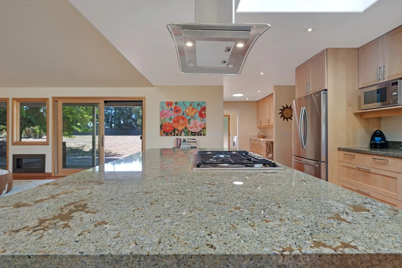 Modern kitchen with a large granite island, built-in gas stove, stainless steel refrigerator, microwave, and a colorful floral painting on the wall. Large sliding glass doors reveal an outdoor area with trees and a hillside.