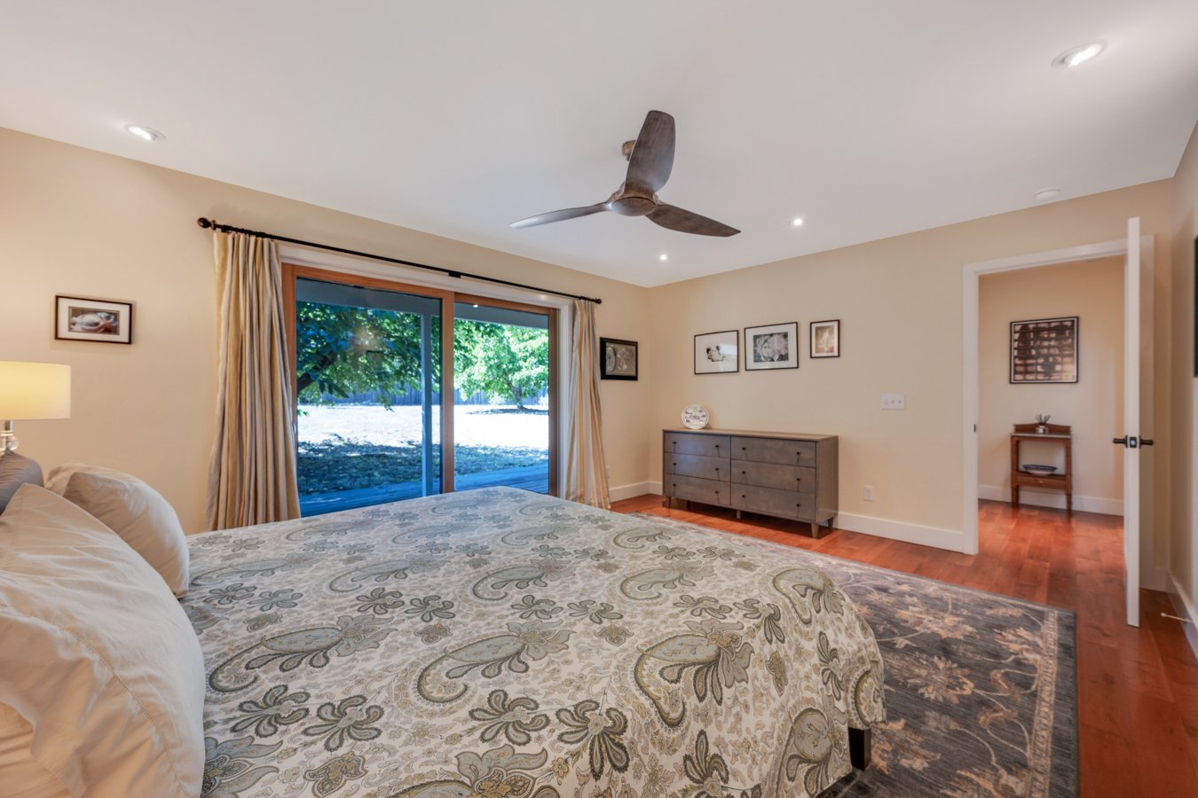 Bedroom with large bed, sliding glass door to backyard, ceiling fan, wooden dresser, framed pictures on walls, hardwood floor, and beige walls.