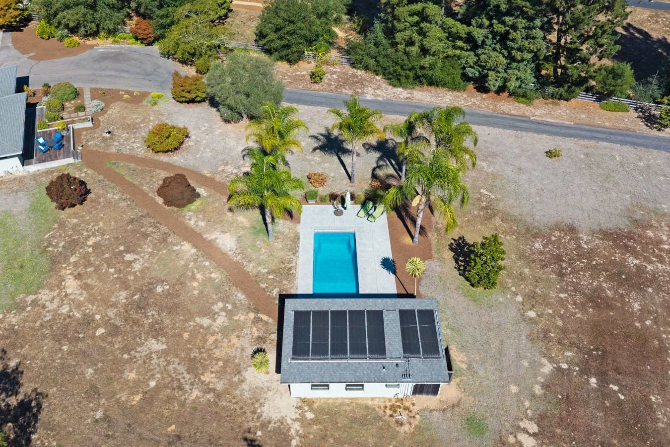 An aerial view of a backyard featuring a small house with solar panels on the roof, a rectangular swimming pool, and several palm trees. The yard has minimal grass, with patches of dirt and some bushes. A road runs along the top of the image, and a n