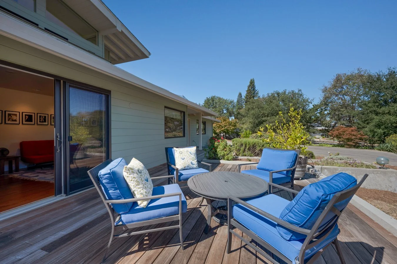 Outdoor patio with four blue cushioned chairs and a round table on a wooden deck, with potted plants and trees in the background, under a clear blue sky.