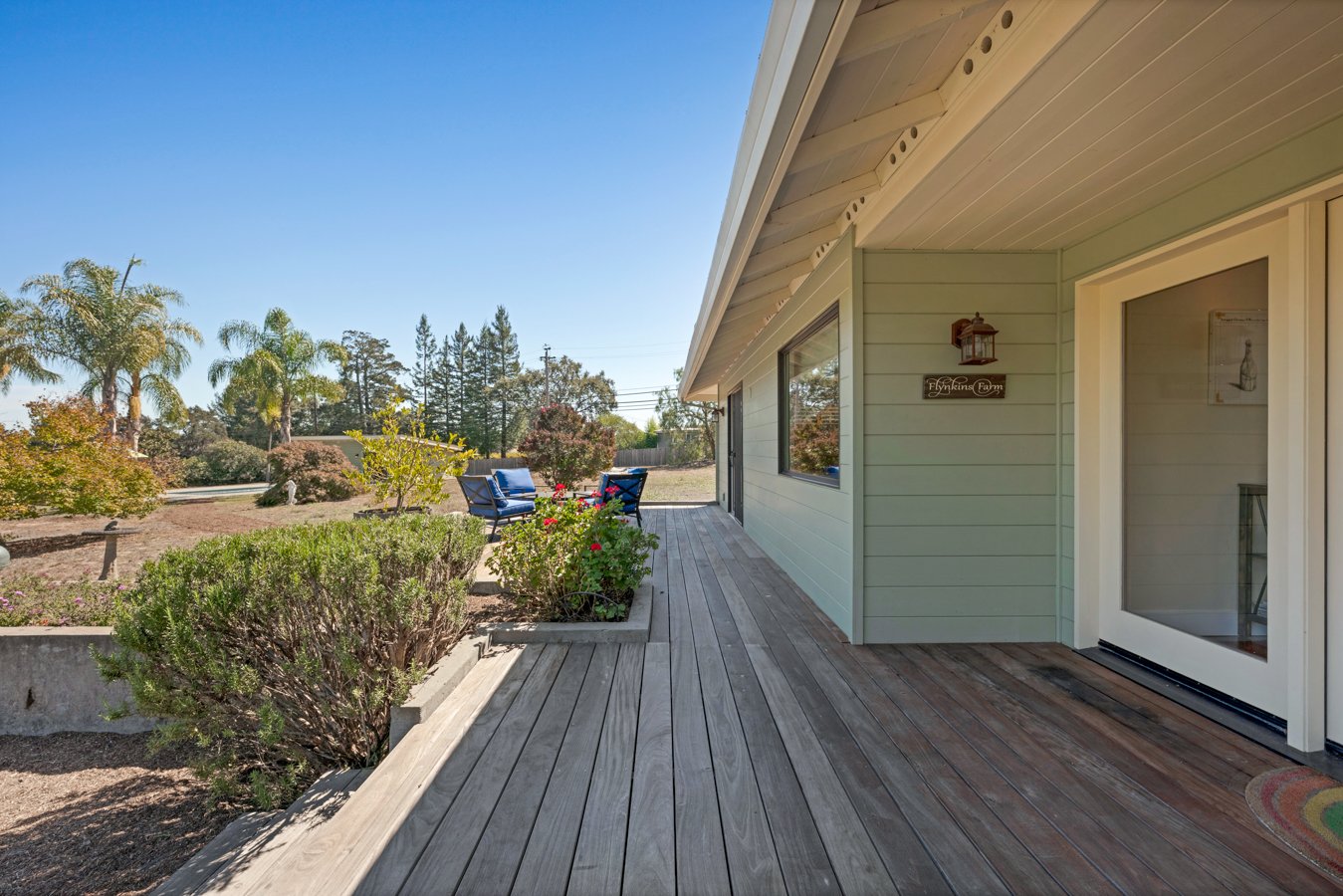 View of a house's wooden porch with outdoor seating and a garden with trees and bushes in the background on a sunny day.