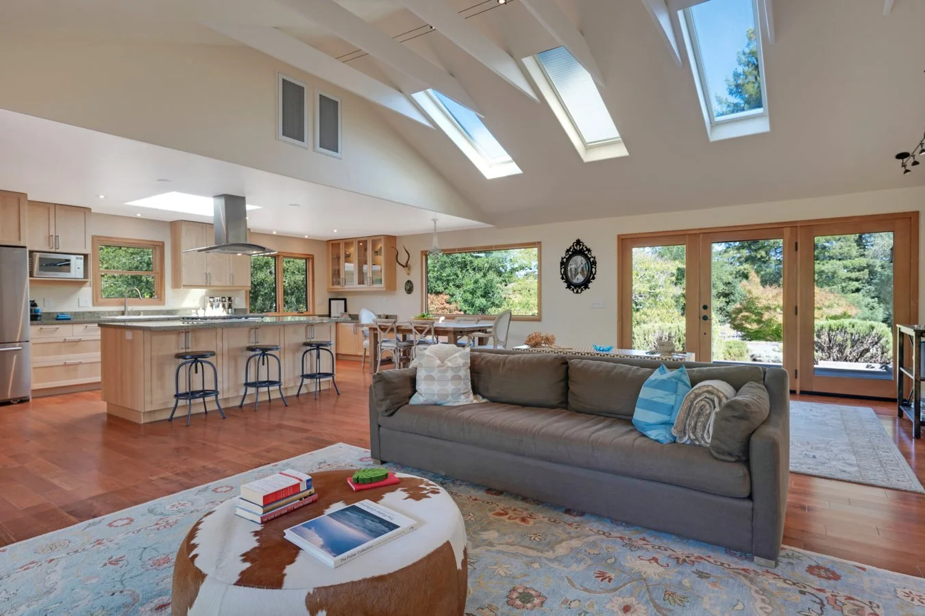 Open-concept living room and kitchen with wood floors, large windows, and skylights, furnished with a gray sofa, a cowhide ottoman, and a dining table, with outdoor greenery visible through the windows and glass doors.