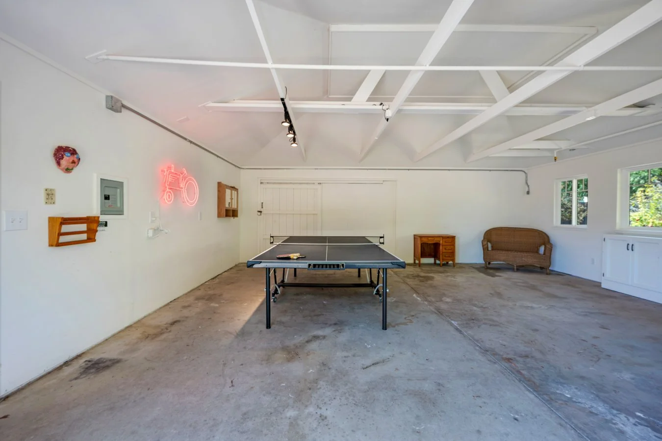 Garage with ping pong table, wicker loveseat, small wooden desk, decorative neon sign, neon face sculpture, picture frame, electrical panel, several small windows, and exposed ceiling beams.