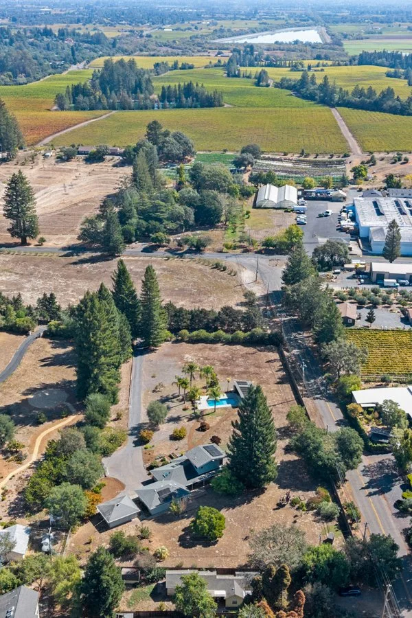 Aerial view of a rural landscape with farmland, trees, a small residential property with a pool, and a commercial building in the distance.