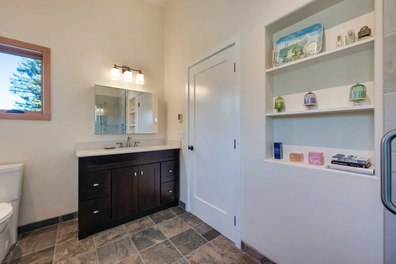 Bathroom with a dark wooden vanity, a mirror with light fixture, a window showing trees outside, and a built-in shelf with decorative items and books.