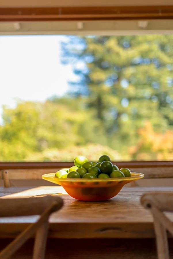 A bowl of green cherries placed on a wooden table near a window with a blurred outdoor view of trees and sky.
