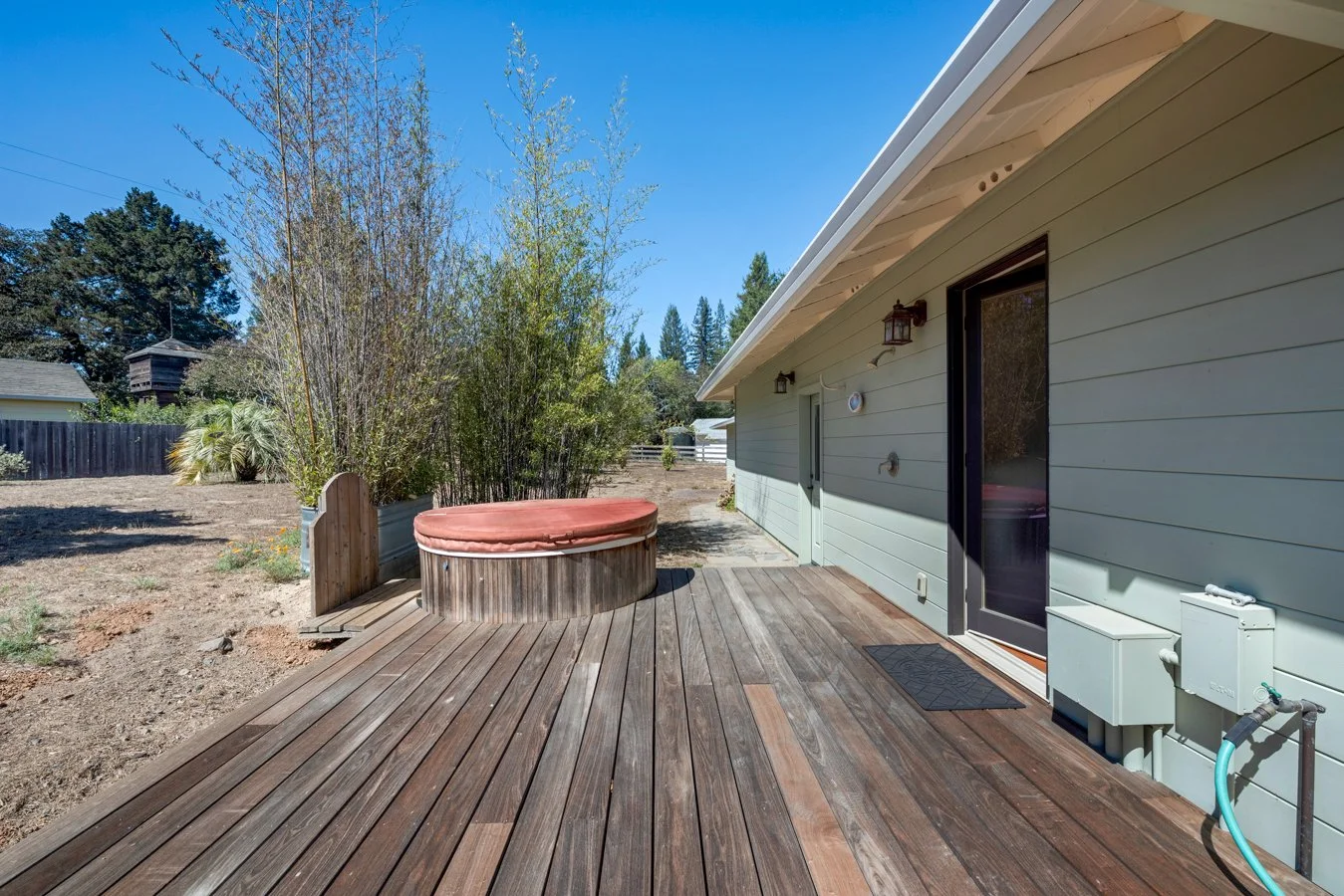 Backyard patio with wooden deck, hot tub with cover, light green house siding, black door with screen, outdoor lighting fixtures, garden hose, surrounding dirt area, plants, trees, and clear blue sky.
