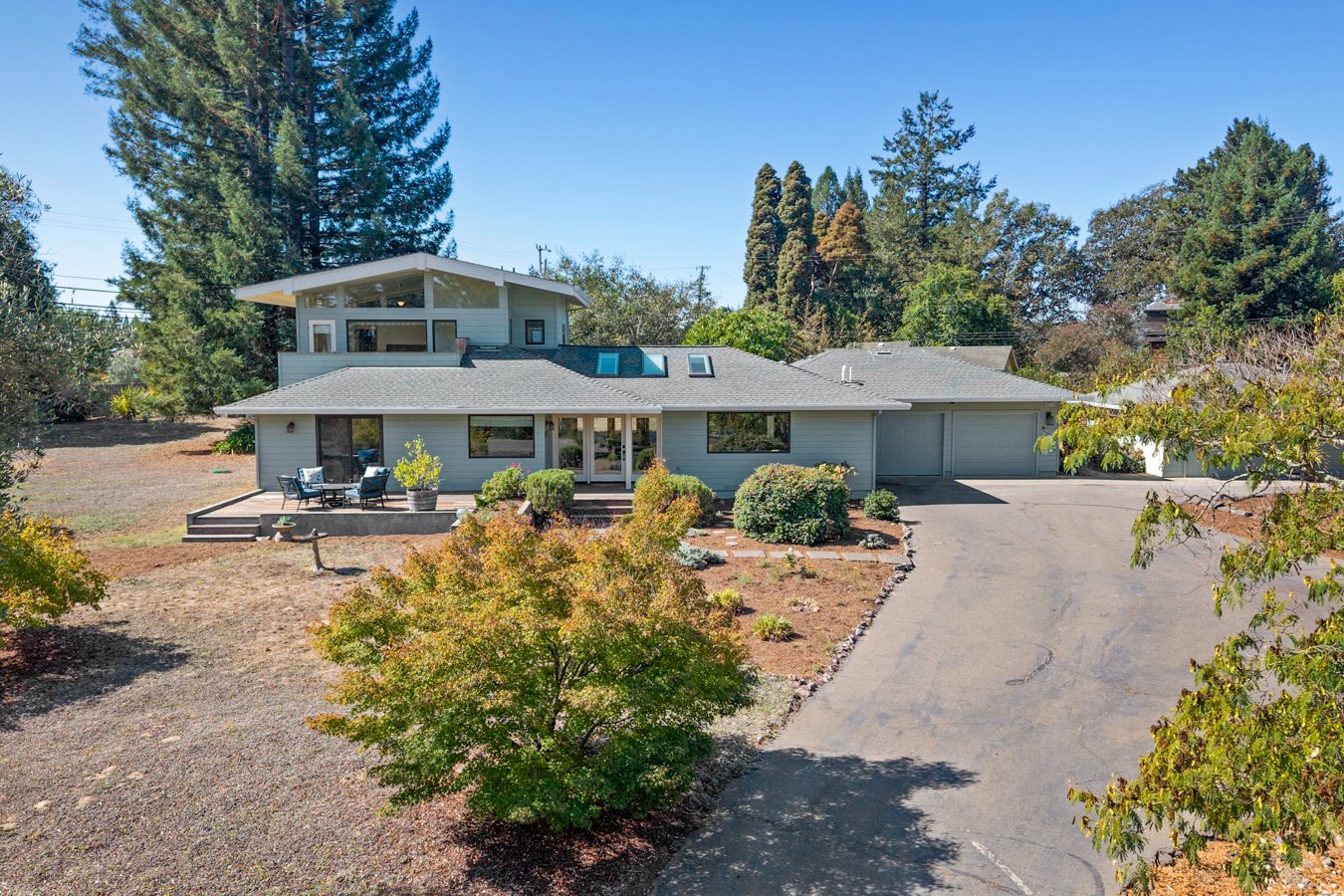 Modern house with multiple roof sections, large windows, and a front porch with outdoor furniture, surrounded by trees and landscaped yard.