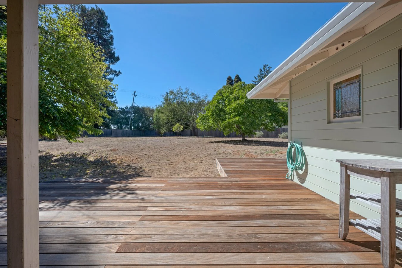 View of a backyard with a wooden deck, a white house wall with a window, and a garden hose. There are several green trees and a clear blue sky.