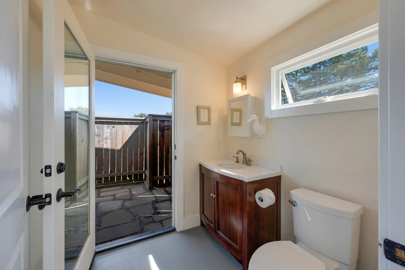 Bathroom with a wooden vanity, a white toilet, a window, and a glass door leading to a small outdoor patio with gray stone flooring and a wooden fence.