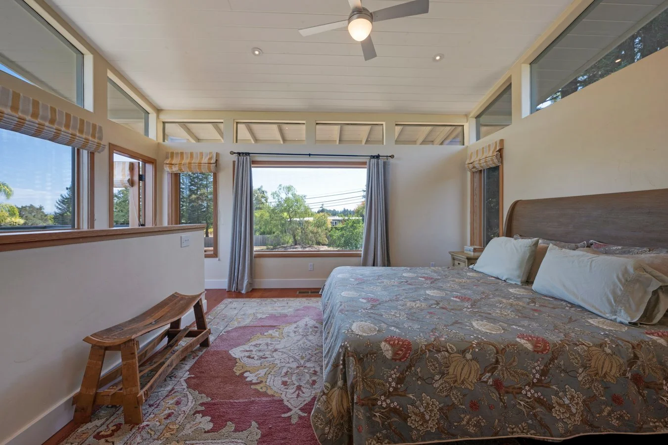 Bright bedroom with a large bed, patterned bedspread, and multiple windows with views of trees and a clear sky, featuring a ceiling fan and light, a bedside table, curtains, and a rug on a wooden floor.