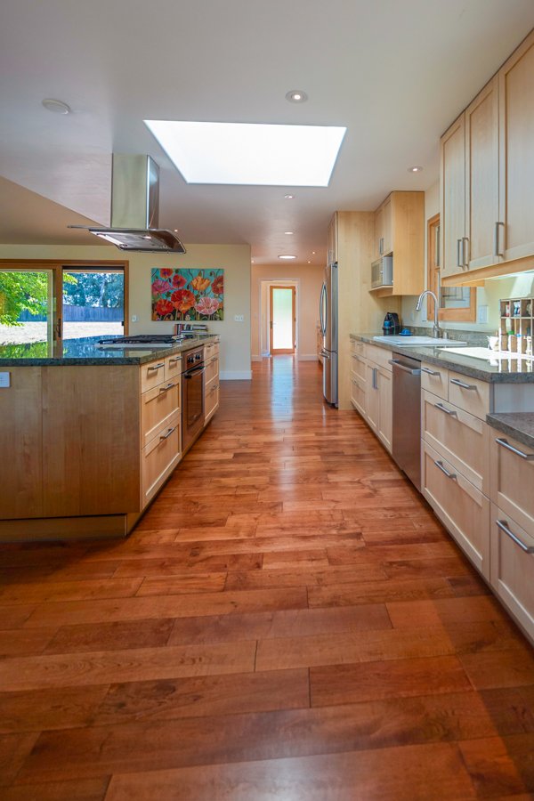 Modern kitchen with hardwood floors, light wood cabinets, stainless steel appliances, a skylight, and a door at the end of the hallway leading outside.