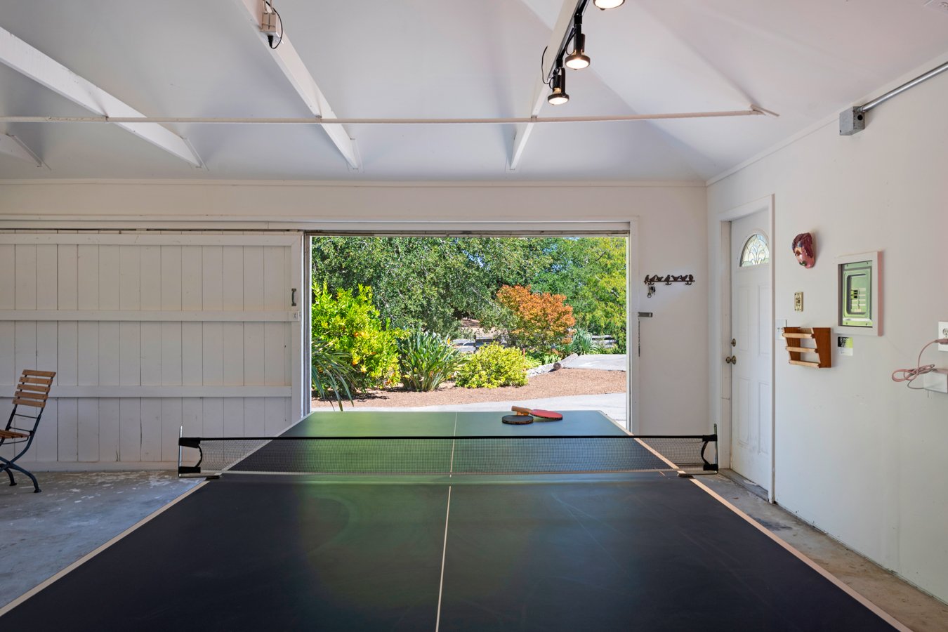 Indoor garage with a ping pong table, paddle and ball, open to a garden outside with trees and plants.