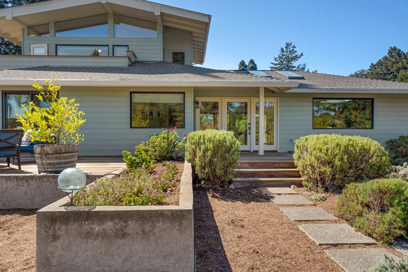 Front view of a modern house with a garden, stepping stones, shrubs, and a patio area under a clear blue sky.