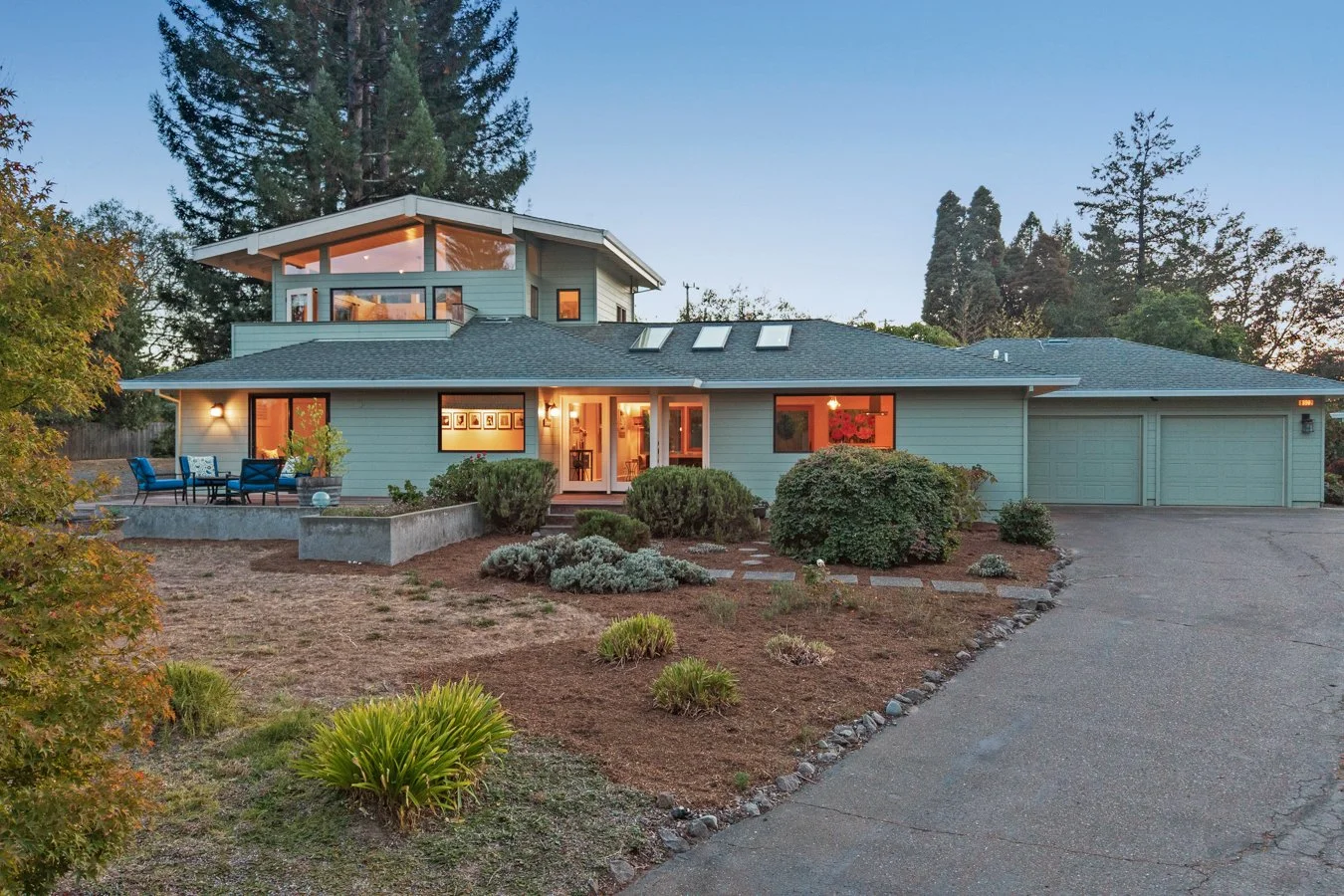 A modern two-story house with green siding, large windows, a driveway, a front lawn, and outdoor seating area at dusk.