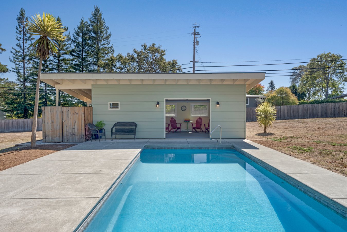 A backyard with a swimming pool, concrete deck, small pool house with patio furniture, trees, and a wooden fence.