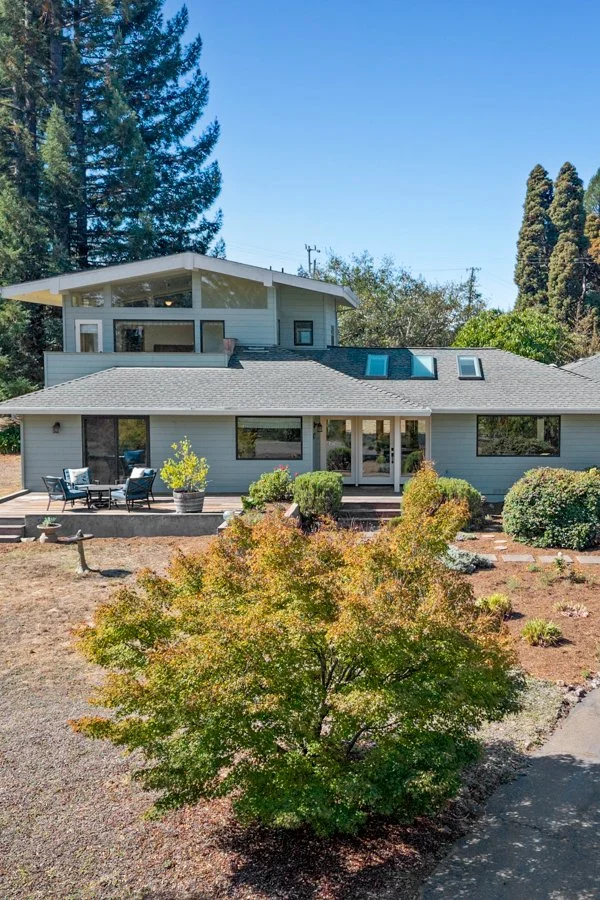 Modern two-story house with a large patio, surrounded by trees and shrubbery on a sunny day.