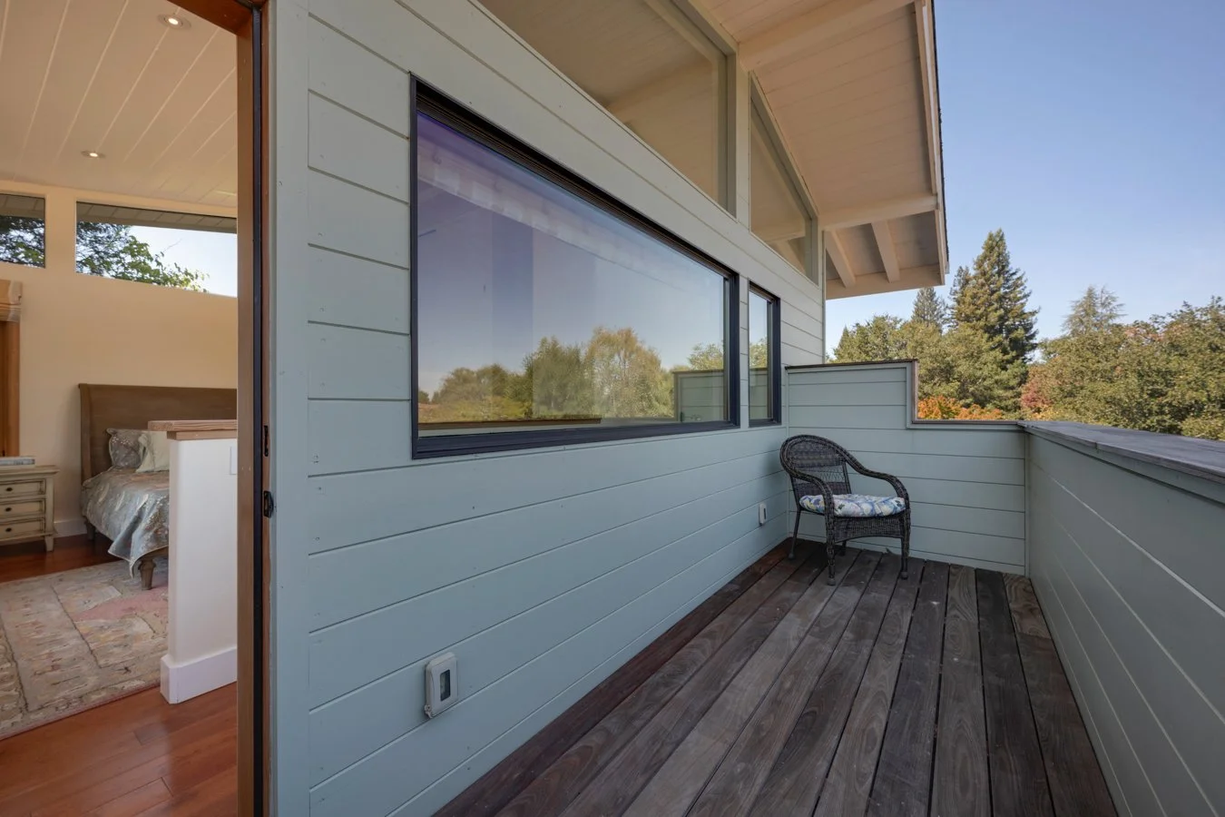 View of a small balcony or porch with a wicker chair and a floral cushion, overlooking trees and a clear blue sky, attached to a home with light-colored siding and large windows.
