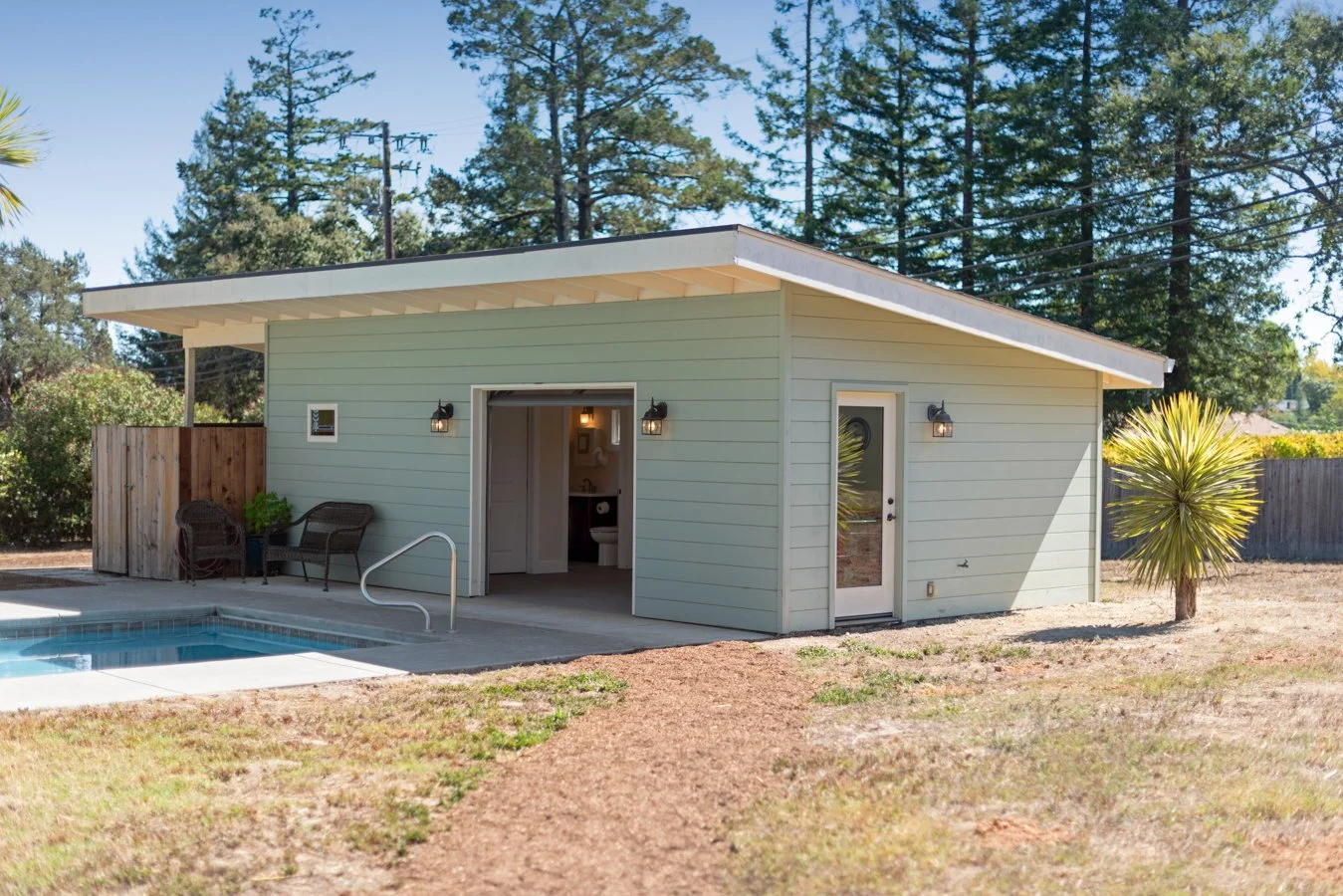 Small house with light green siding, open door, outdoor seating, and a small pool in the yard, surrounded by trees and a wooden fence.