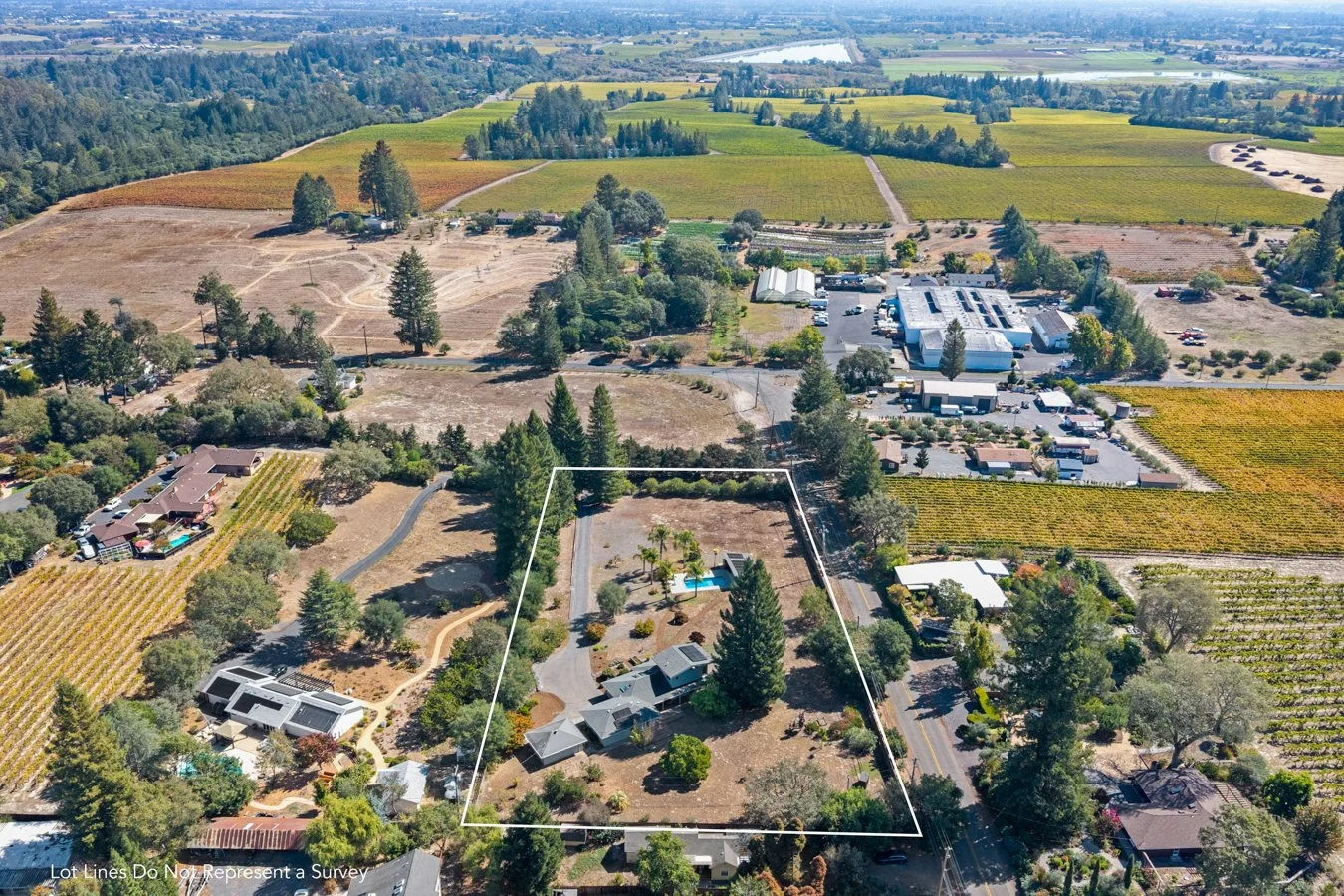 Aerial view of a rural property outlined in white boundary lines, featuring a house with a swimming pool, surrounded by trees, gardens, and nearby roads, with neighboring properties and agricultural fields in the background.
