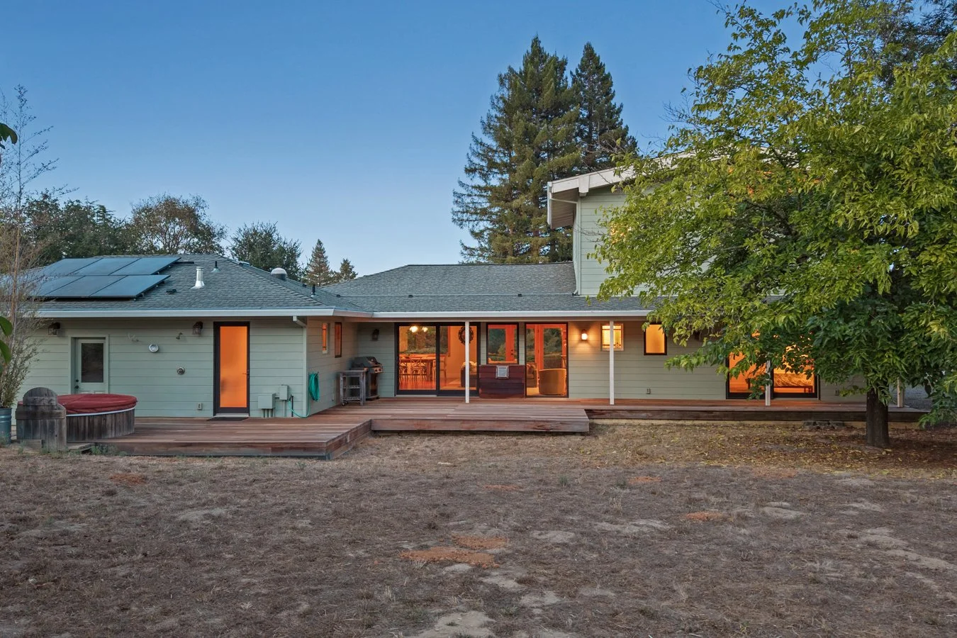 Rear view of a house with a wooden deck, large sliding glass doors, a hot tub, a barbecue grill, and a tree in the backyard, during dusk.
